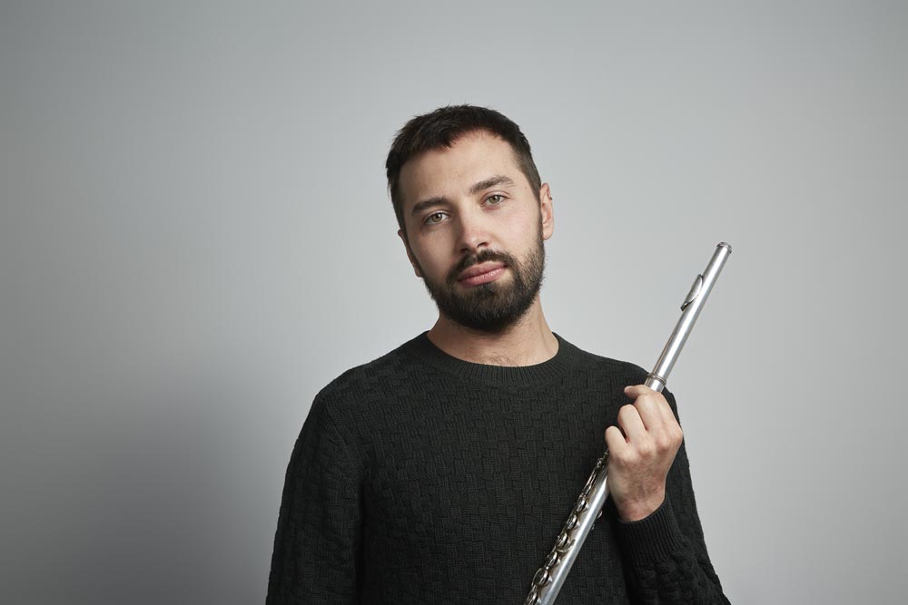 A man wearing a black jumper holding a flute and looking at the camera