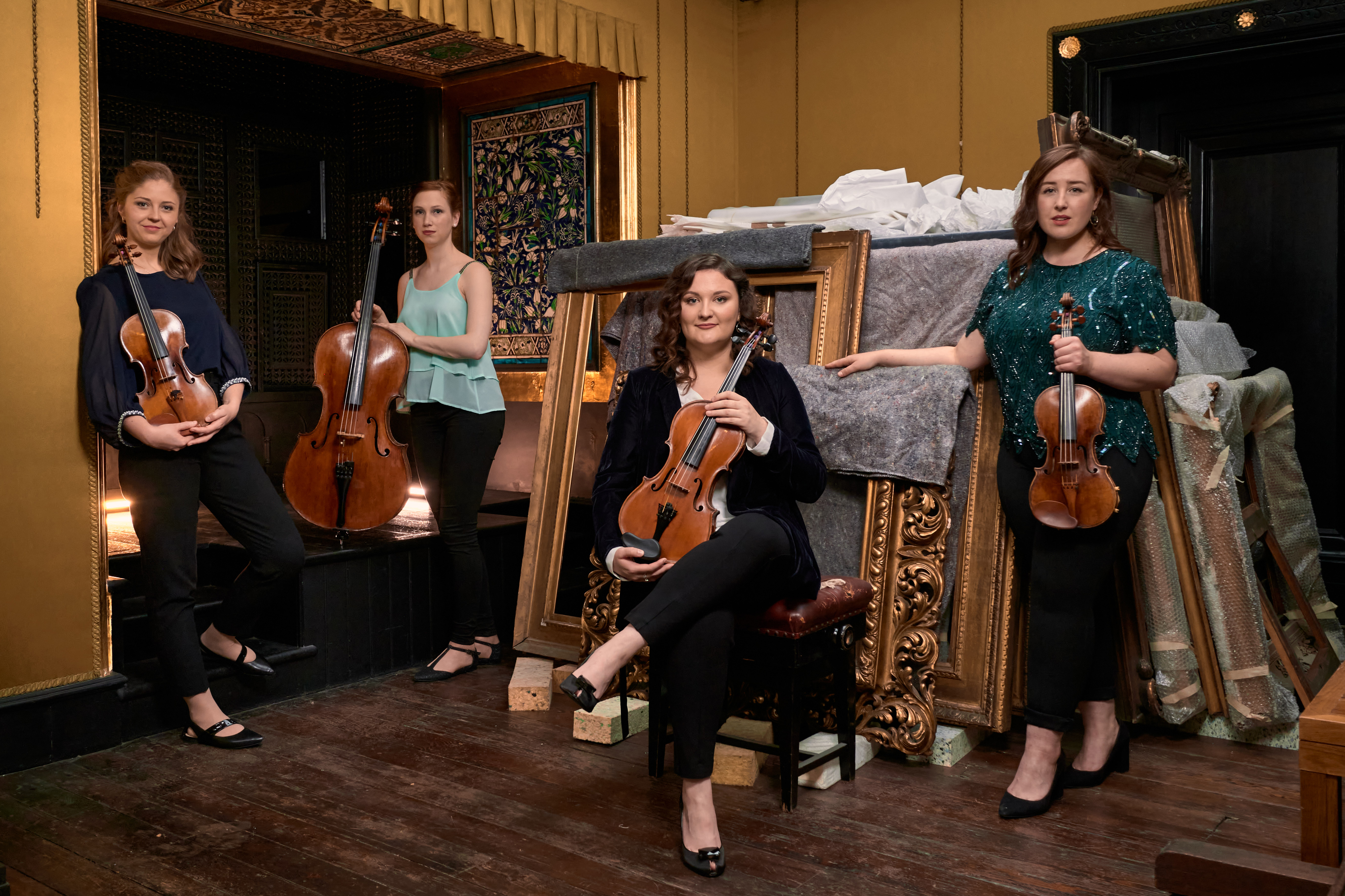 A string quartet standing in an ornate room in front of picture frames holding their instruments