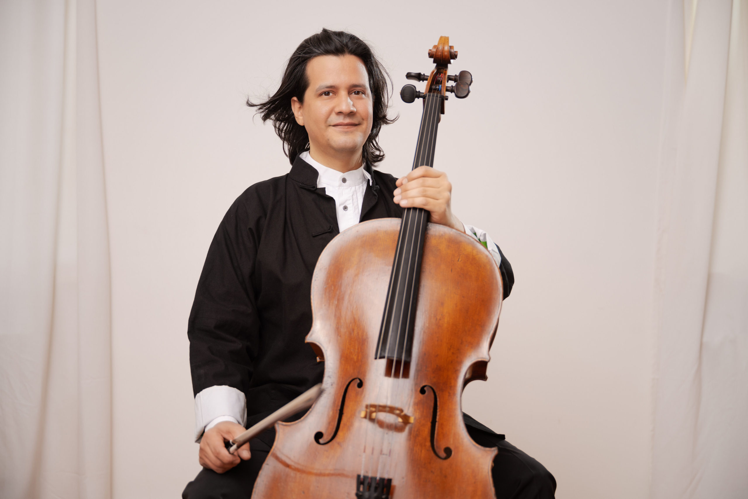 Claudio with a white background, holding his cello and looking at the camera