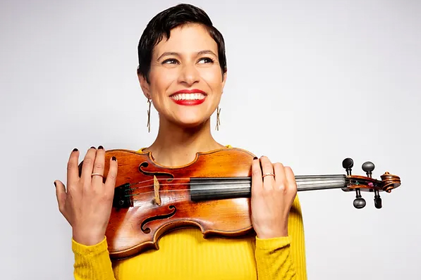 A woman with short hair wearing yellow, holding a violin and smiling