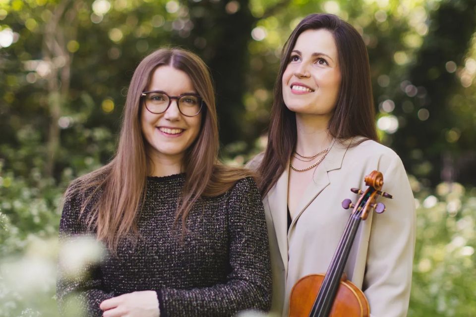Two young women stand outside and smile holding a violin