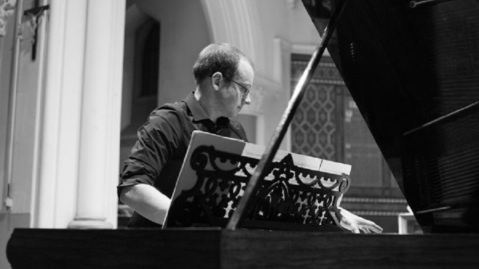 A black and white photo of male alumna, wearing glasses, wearing a dark shirt, performing on a piano.
