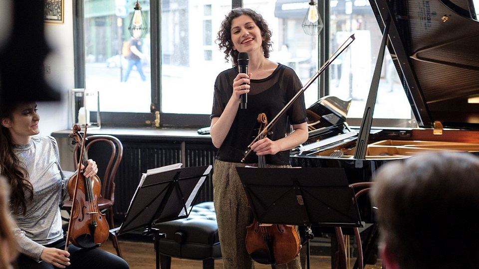 Female alumna, holding a violin and a microphone, standing in front of an audience and next to a piano.