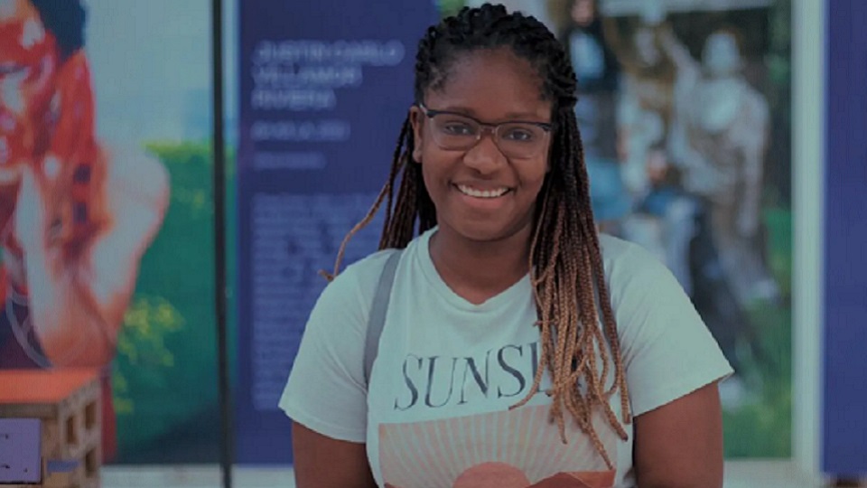 A black female alumni, wearing a light coloured t-shirt looking directly at the camera and smiling