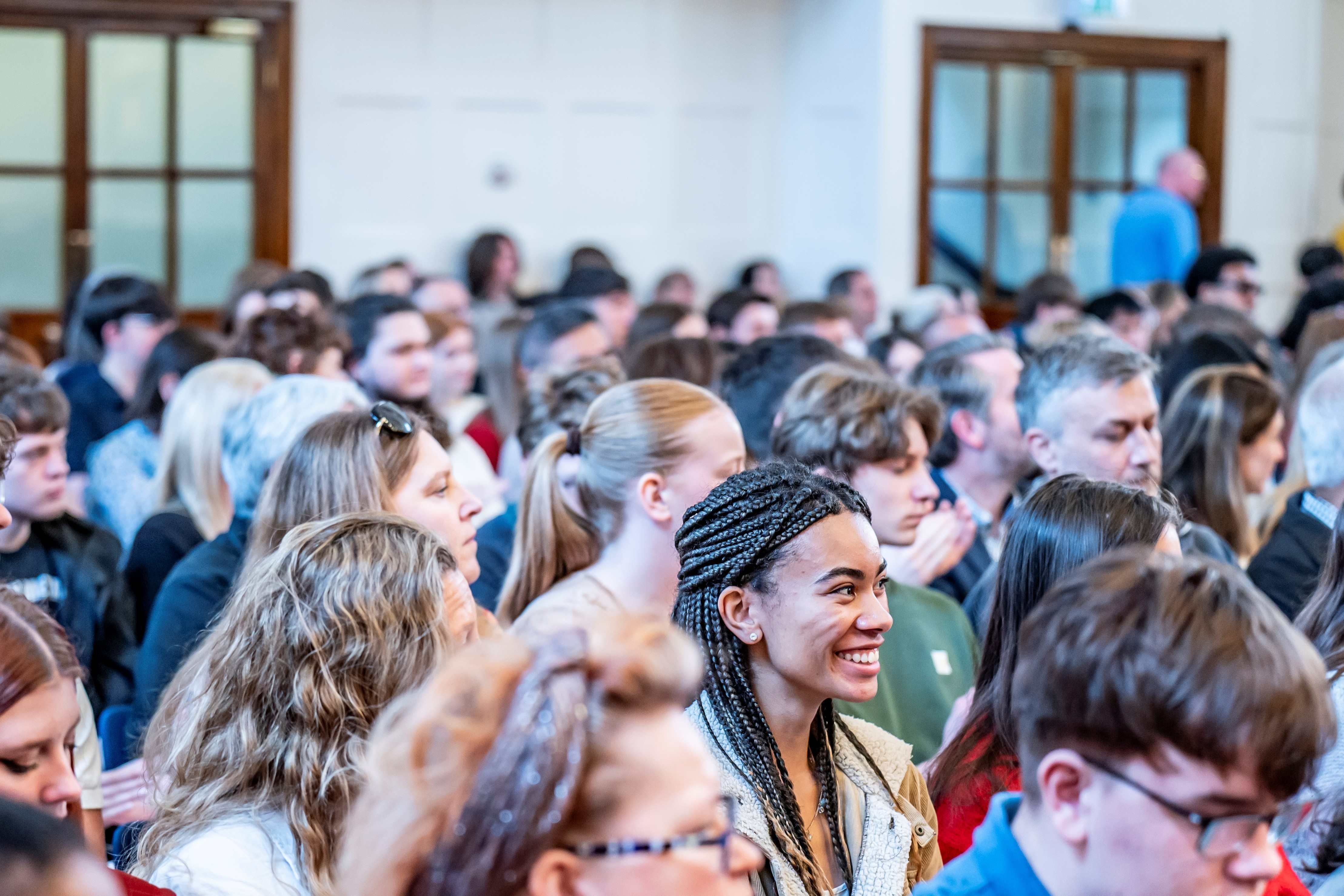 A large audience is seated closely together in a bright event hall, facing toward the front. In the foreground, a female individual with long, dark braided hair, wearing a light textured sweater and a jacket, appears to be smiling as they listen. They are surrounded by other attendees, some of whom are clapping. The room has tall windows and wooden-framed doors in the background. A large audience is seated closely together in a bright event hall, facing toward the front. In the foreground, a female individual with long, dark braided hair, wearing a light textured sweater and a jacket, appears to be smiling as they listen. They are surrounded by other attendees, some of whom are clapping. The room has tall windows and wooden-framed doors in the background.
