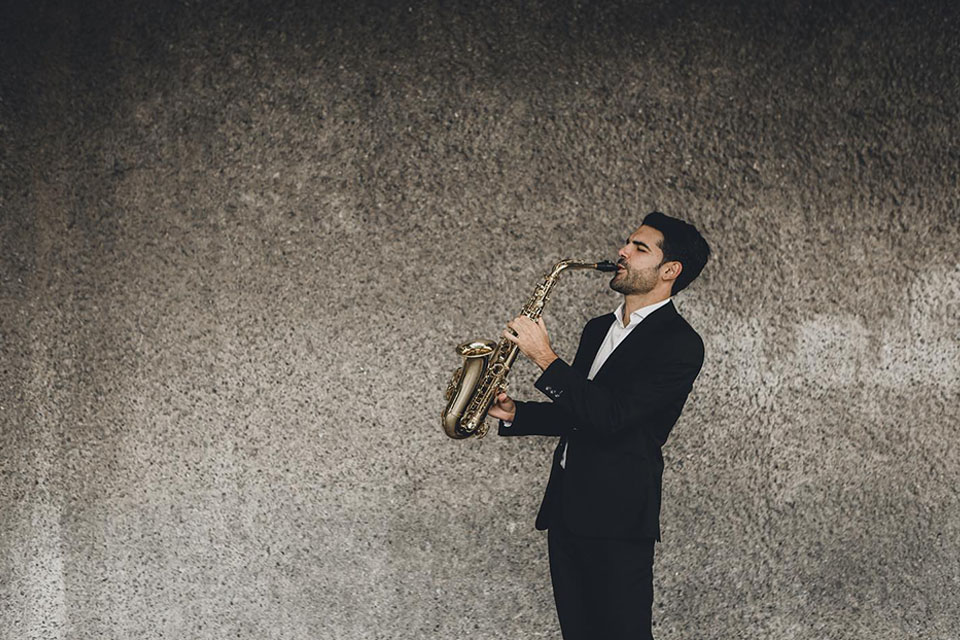 Manu Brazo holds saxophone in front of a grey backdrop