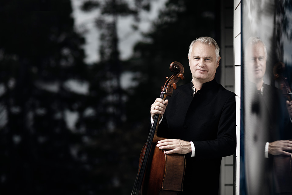 Torleif Thedeen stands outside next to some mirrored glass, holding a cello