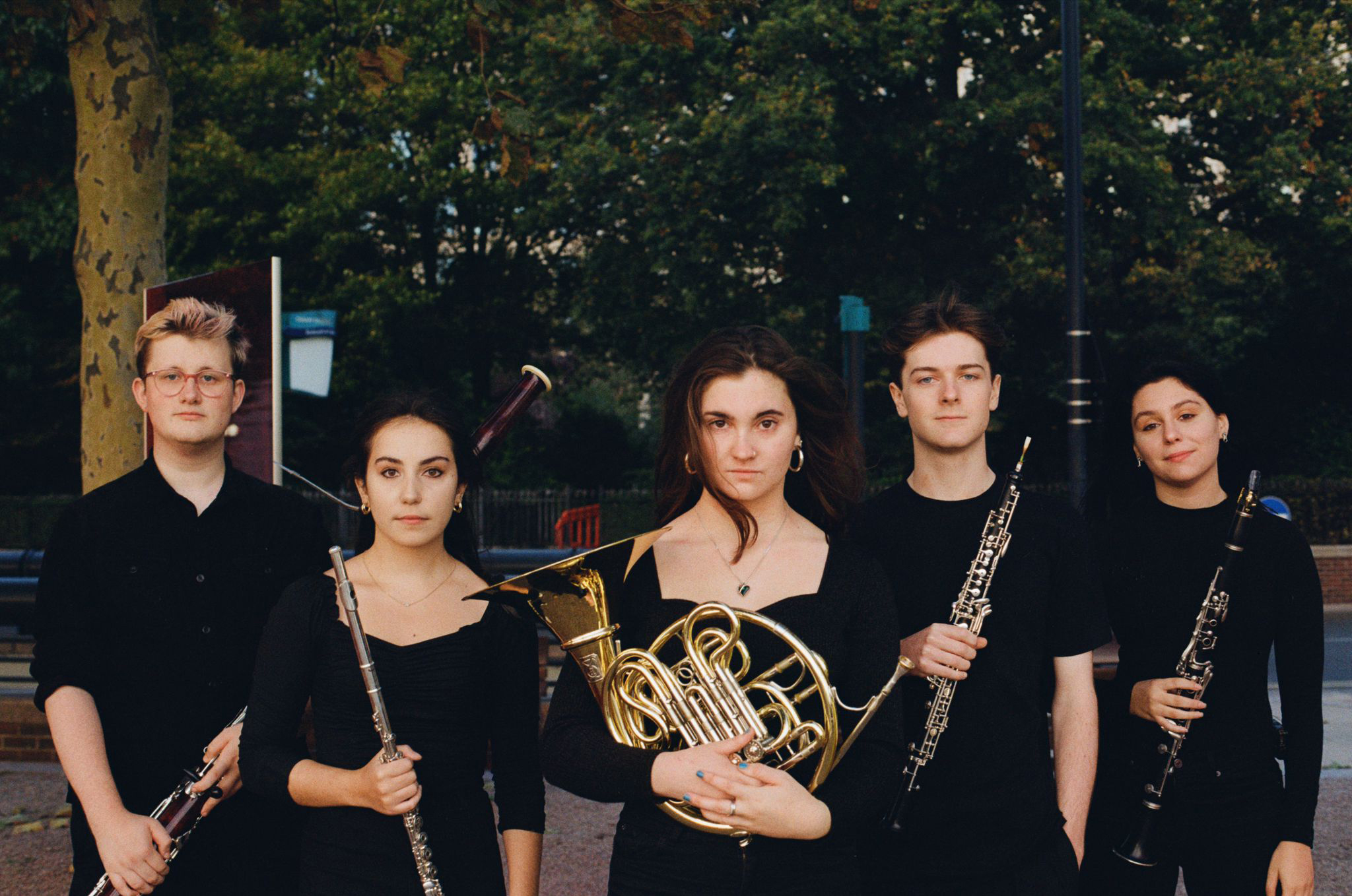A photograph of the five members of Aeolian Winds wearing black, standing outside, holding their instruments and looking at the camera