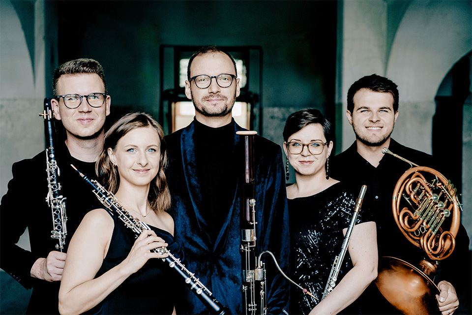 Five woodwind musicians smiling stood holding instruments in an old building