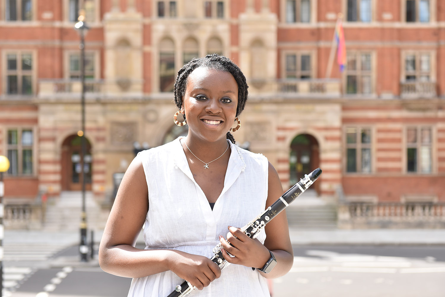A girl smiling wearing a white dress stands in front of the RCM holding a clarinet