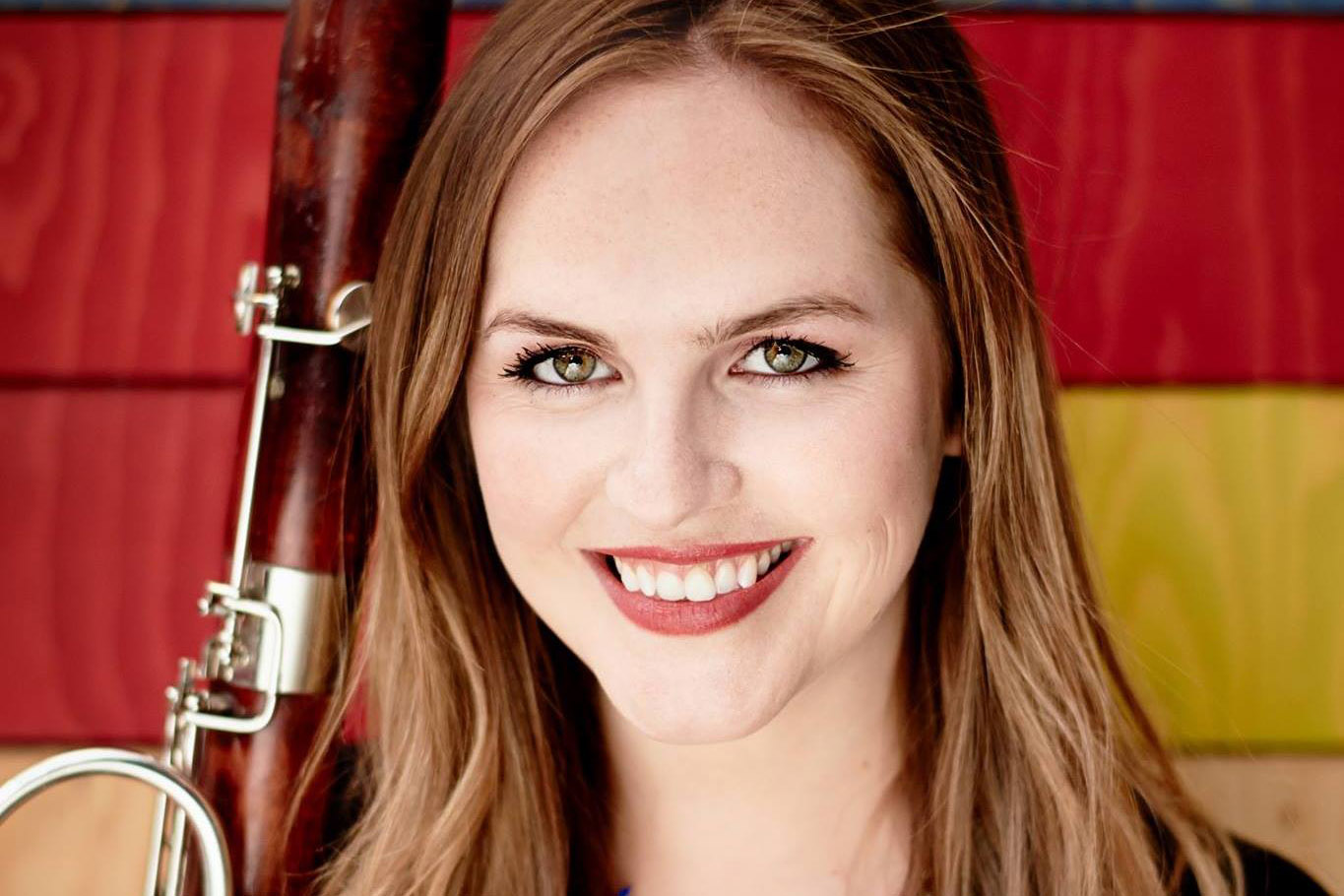 A close up image of a young woman smiling at the camera holding an oboe in front of a colourful background