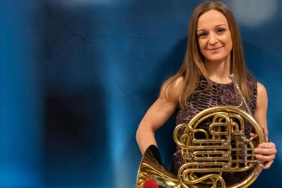 A woman standing in front of a blue background holding a French horn