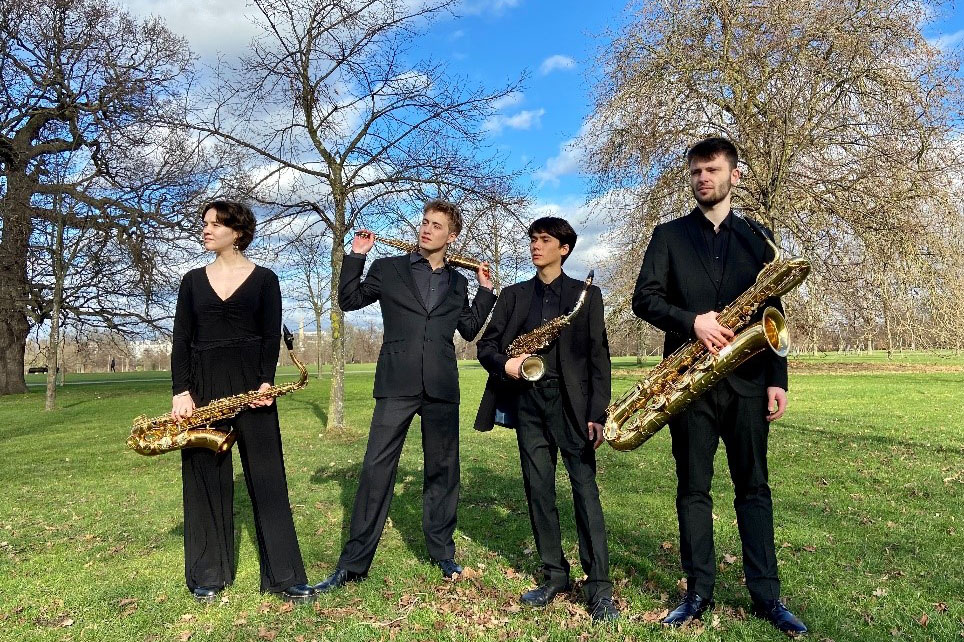 Four people holding saxophones, wearing black standing in a grassy park with blue sky and winter trees