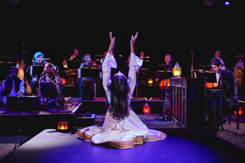 A woman wearing a white dress kneels on stage with her hands in the air looking up, in front of a group of musicians