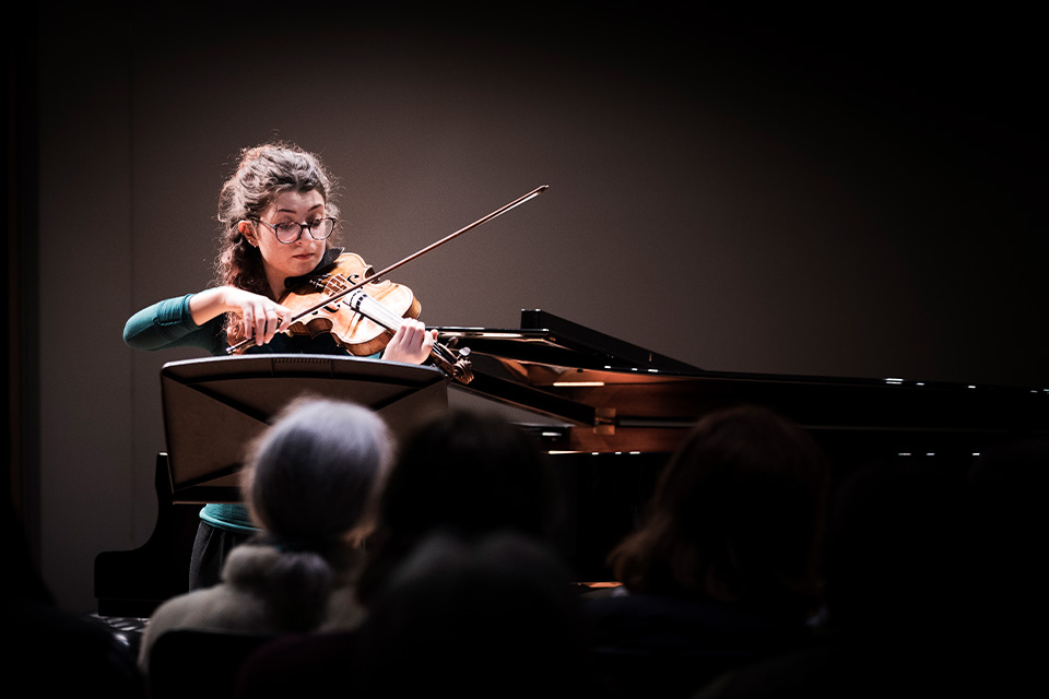 A violinist performs in a dark room in front of an audience