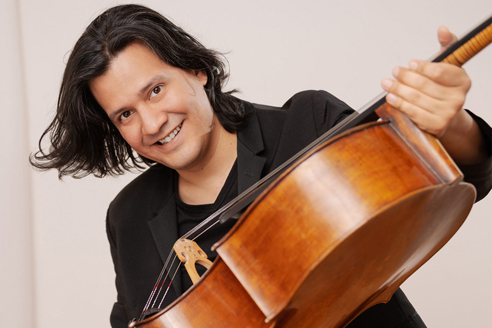 Claudio Bohórque, wearing black, holds his cello up and smiles