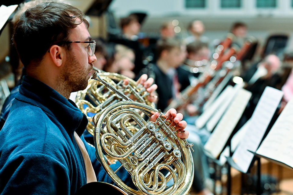 A man in a blue shirt plays the horn with an orchestra