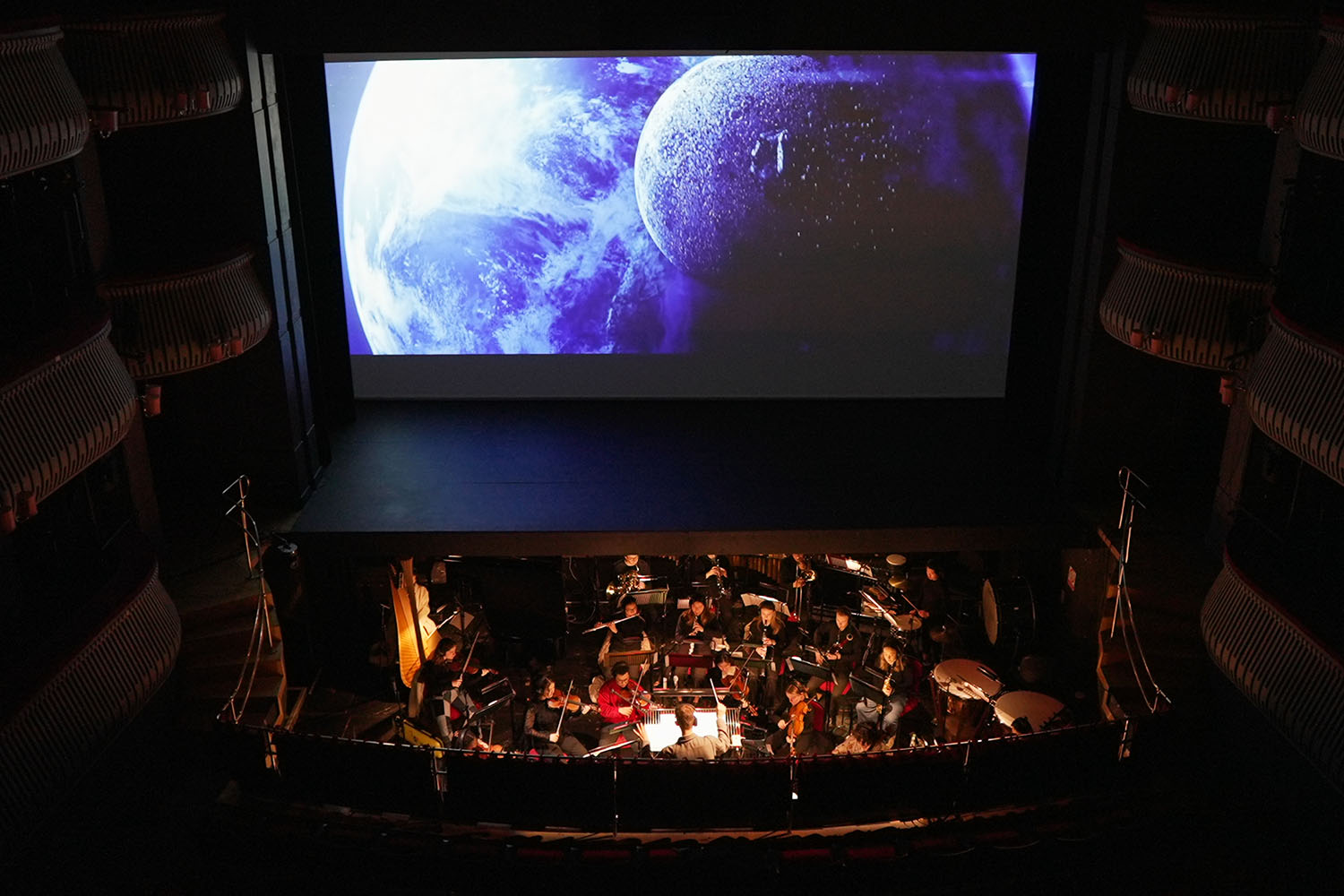 The orchestra in the Britten Theatre below a screen on stage showing the moon and Earth