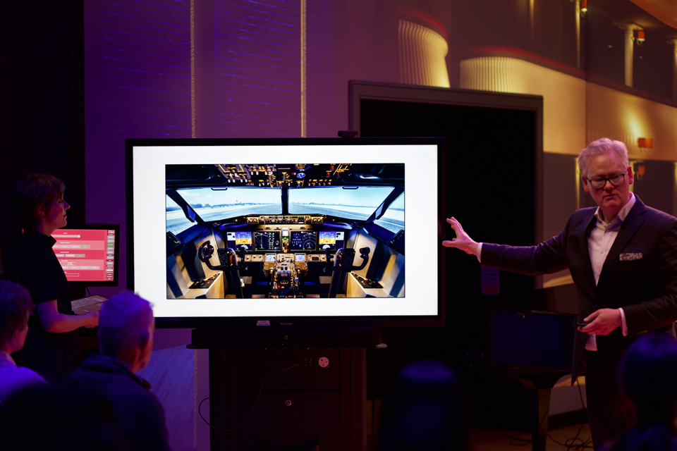 A man gestures at an image of an aeroplane cockpit on a screen