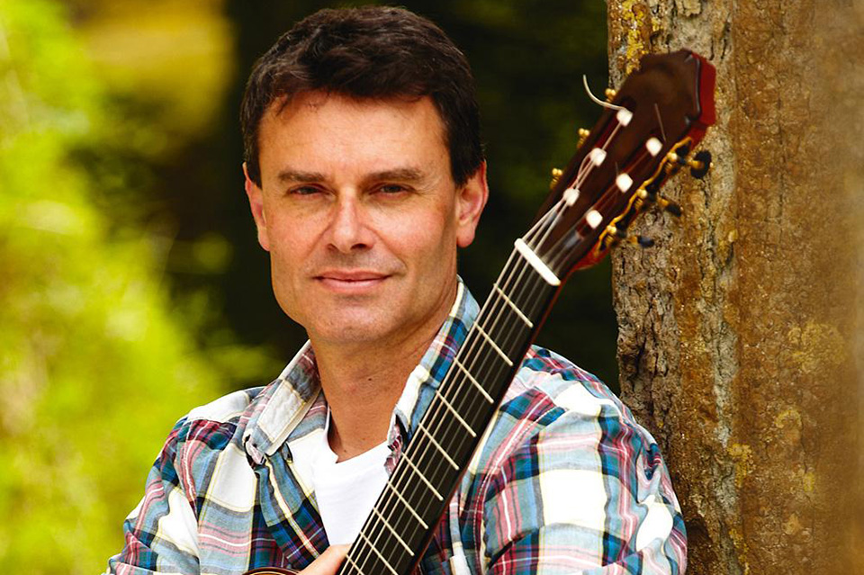 Craig Ogden is pictured outdoors, sat against a tree, holding his guitar