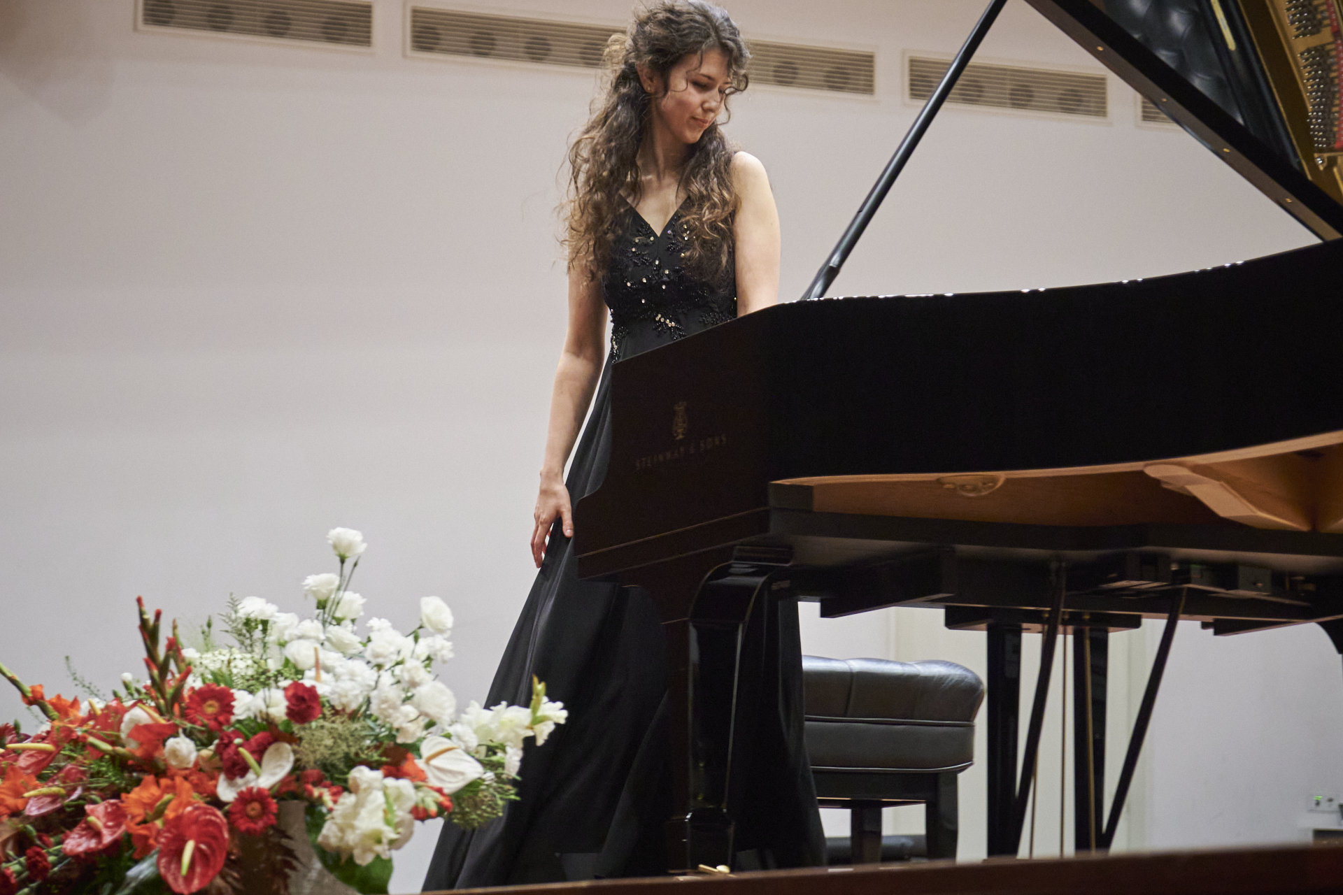 A lady in a long black dress looking down at a grand piano, with flowers in the foreground