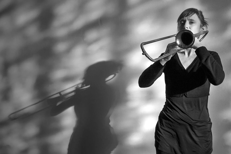 A black and white photo of Emily White playing the trombone, her shadow projected just behind her