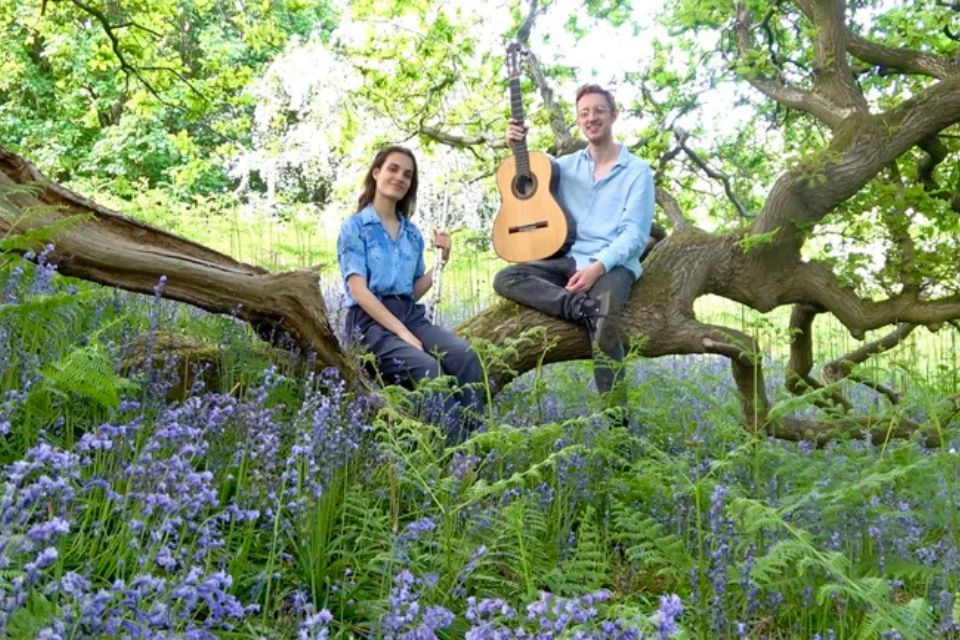 A man holding a guitar and woman holding a flute sit on a tree branch surrounded by purple flowers