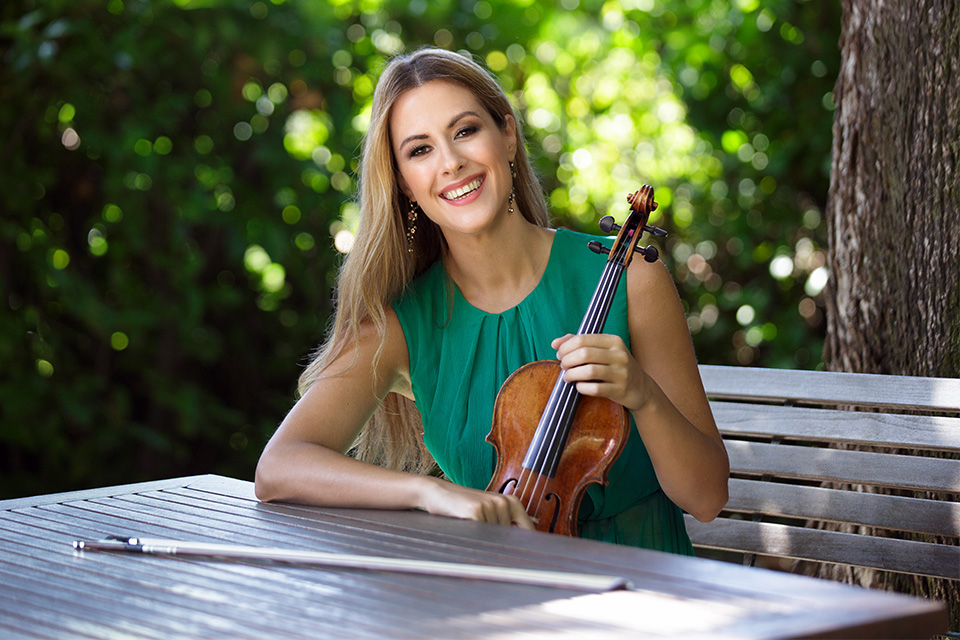 Francesca Dego, wearing a green dress, holds her violin whilst sitting at an outdoor table and smiles