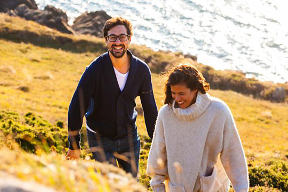 A man and woman laugh as they walk through a field, the sea behind them