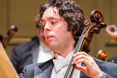 A man with curly hair wearing a tuxedo performing cello on stage