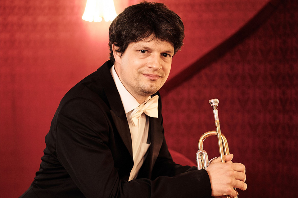 Immanuel Richter, wearing a suit and bow tie and standing against a red background, looks to camera