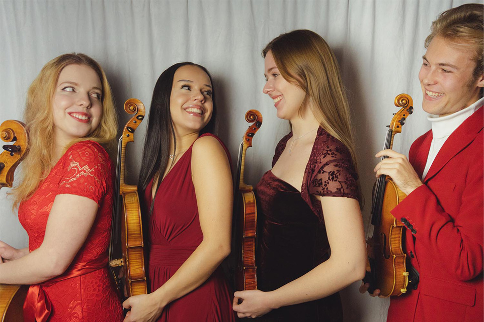 String quartet musicians in a line wearing red holding their instruments and smiling at each other