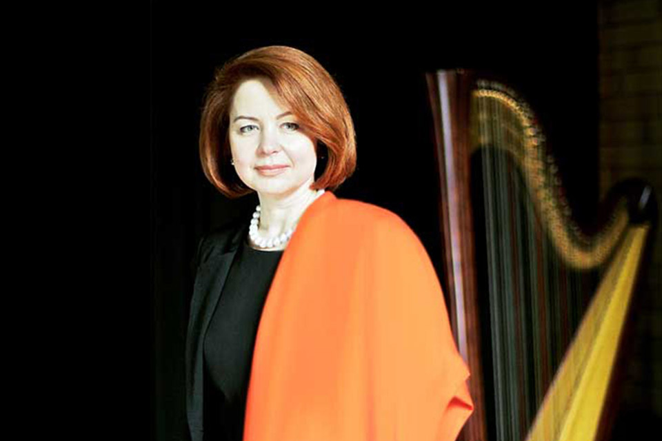 A woman wearing black and orange stands in front of a harp against a black backdrop