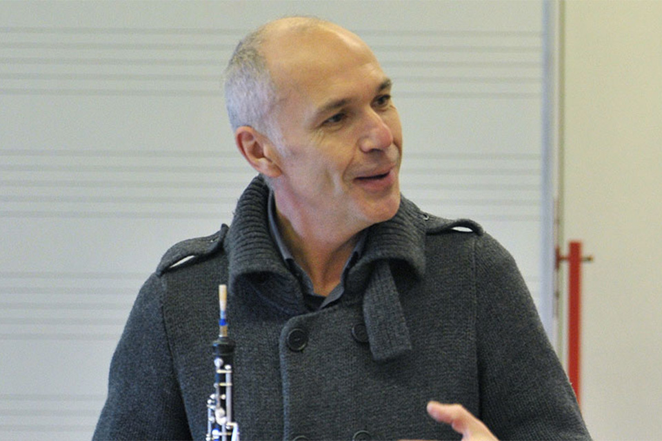 Jacques Tys, wearing a grey coat, holds his oboe and is speaking
