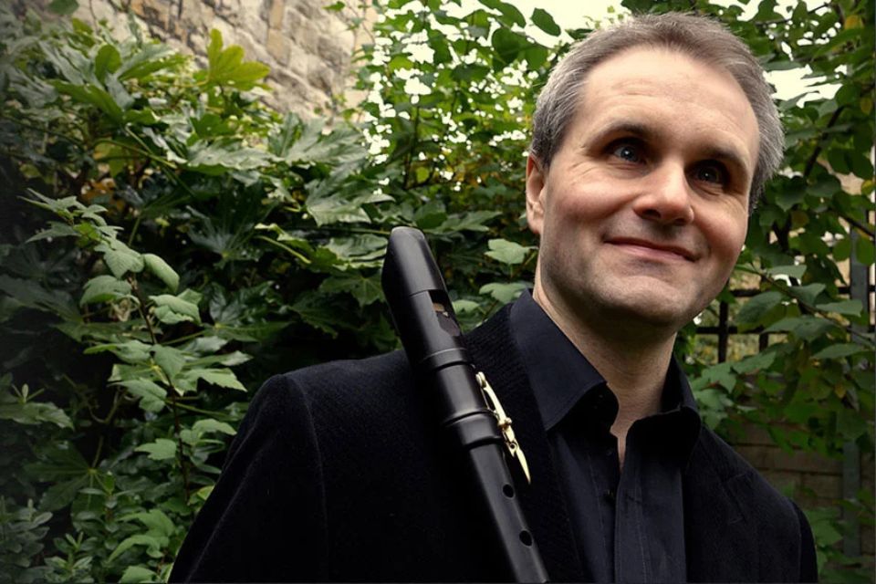 James Risdon wears a black shirt and holds his recorder in front of a leafy background