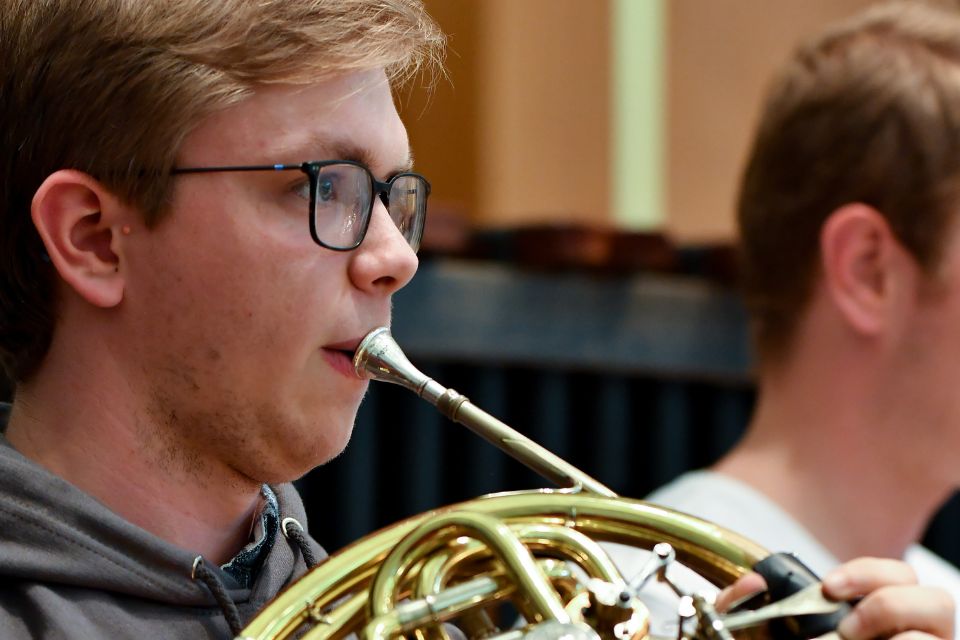 A young boy with blonde hair and glasses plays the horn
