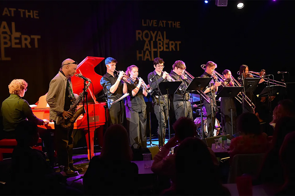 A group of young people play instruments on stage with a projection behind that says 'Live at the Royal Albert Hall'