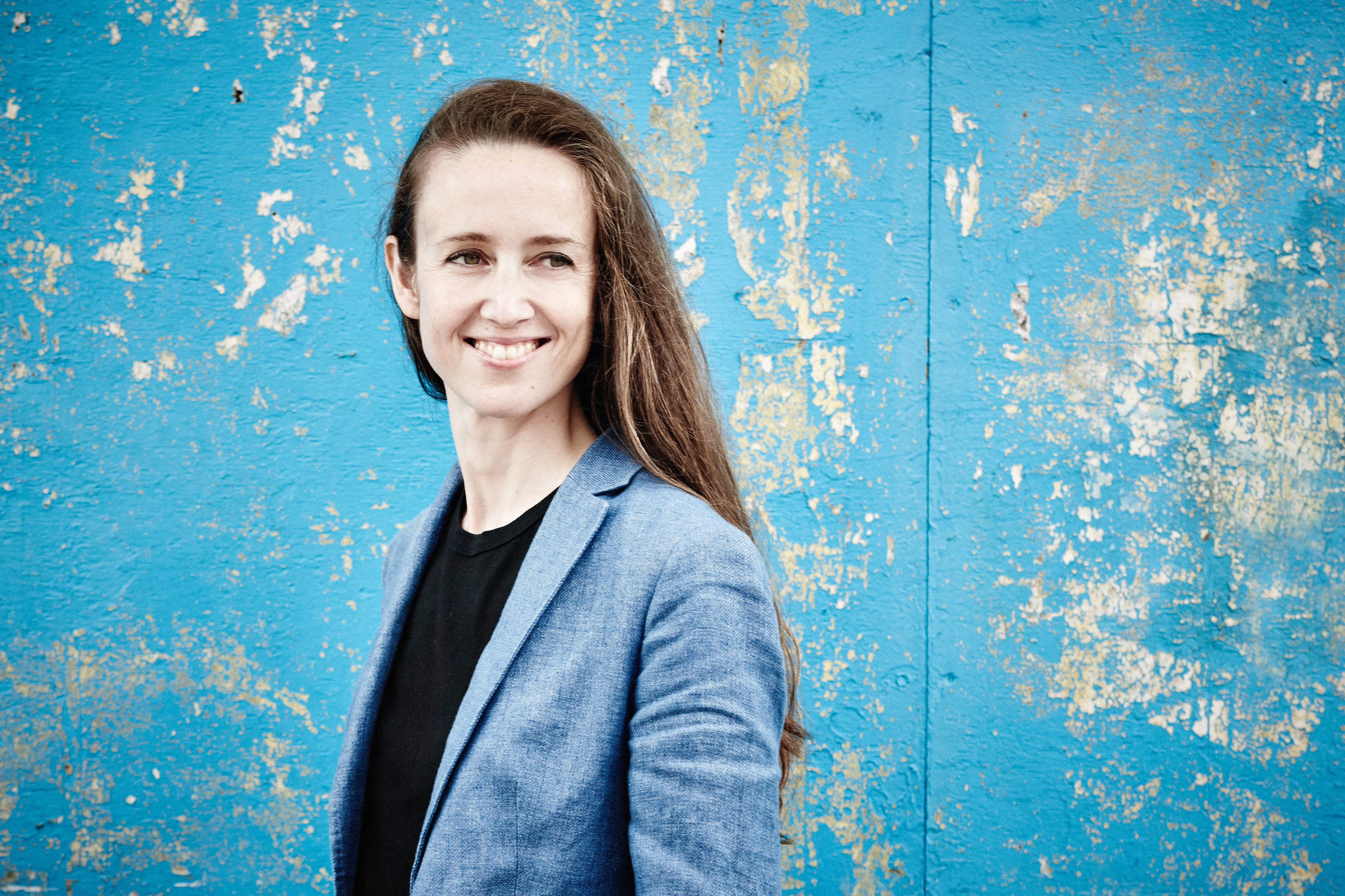 A portrait of conductor Jessica Cottis smiling against a painted blue wall