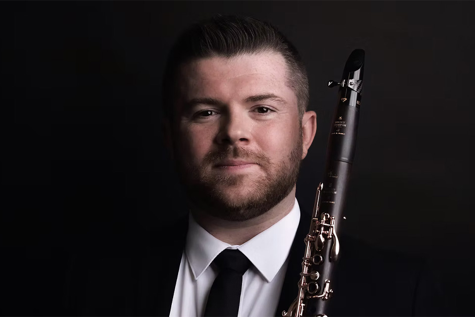 Julian Bliss wearing a suit and tie, and holding his clarinet, smiles at the camera