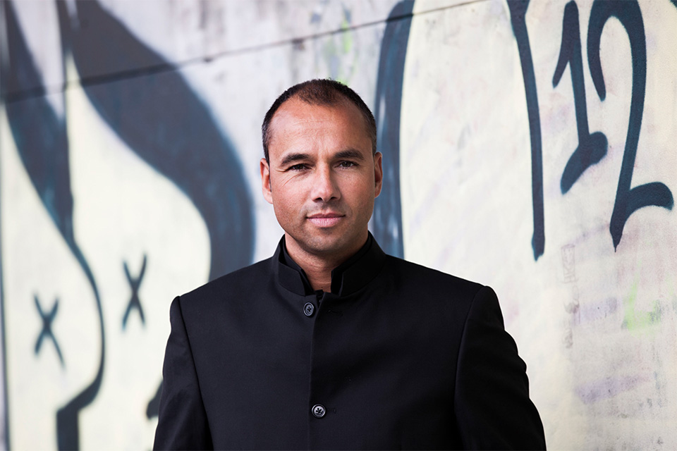 Lawrence Renes wears a black jacket and stands in front of a patterned wall