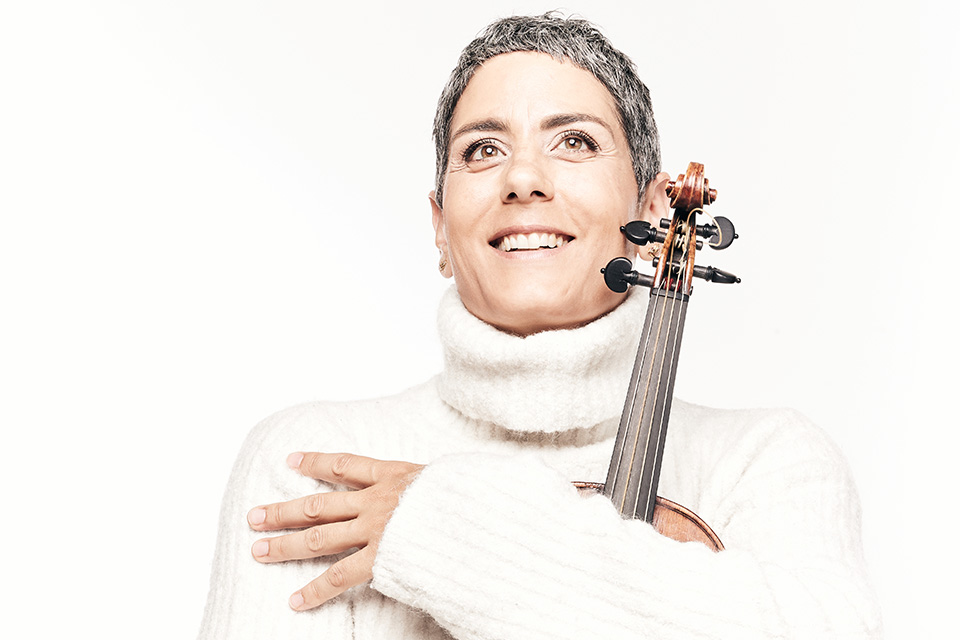 Leila Schayegh, wearing white and standing against a white background, hugs her violin to her