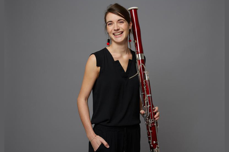 Lola Descours, wearing black and standing against a grey background, holds her bassoon and smiles