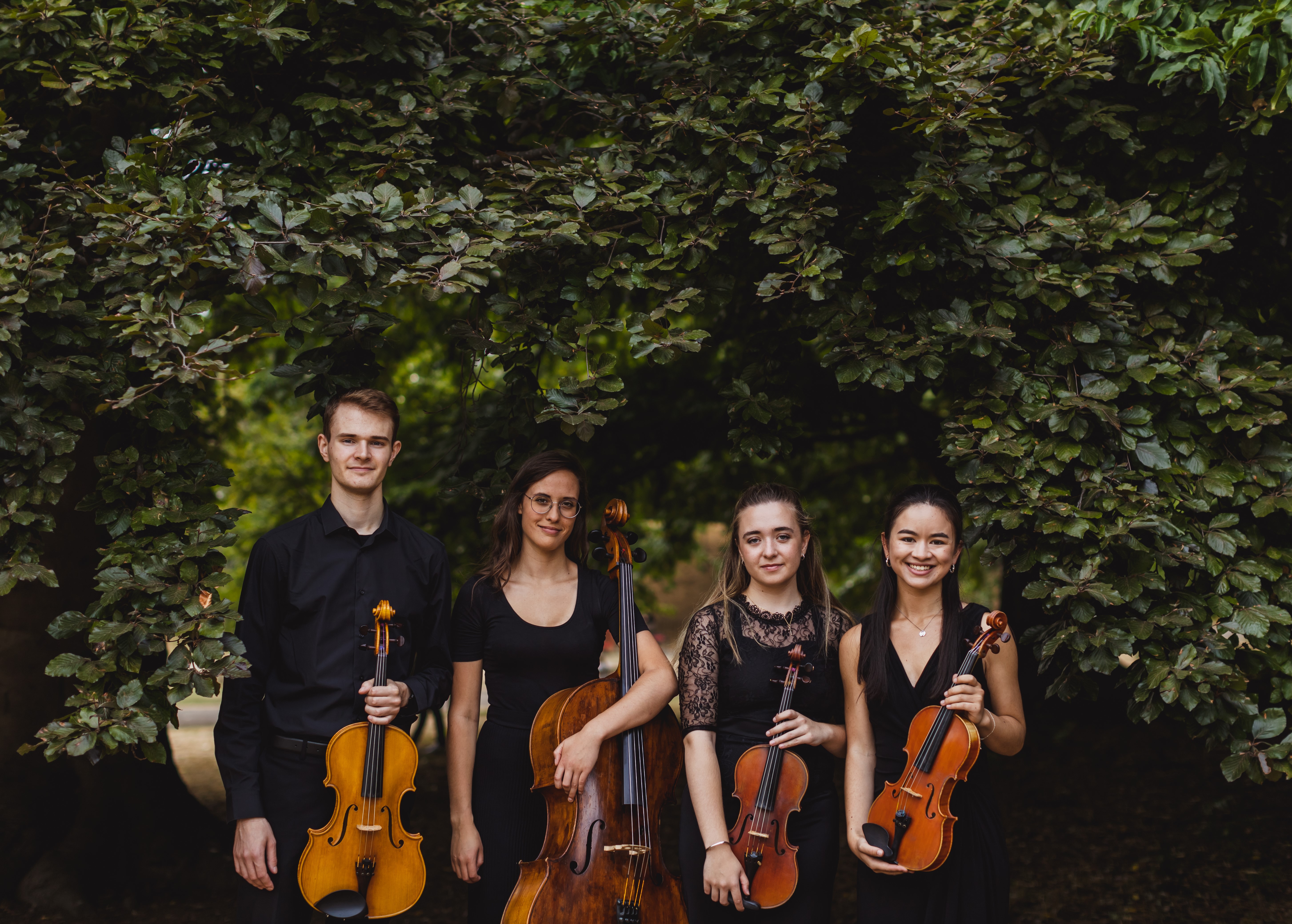 Morassi Quartet sitting on a bench and holding their string instruments smiling with bushes and greenery behind them
