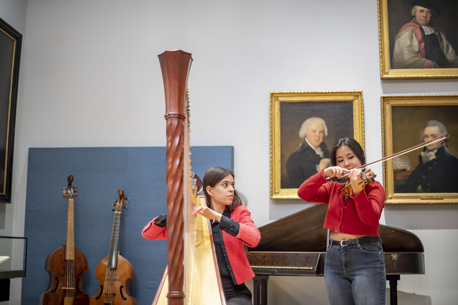 A violinist and a harpist performing against a backdrop of paintings and old instruments in the Royal College of Music Museum