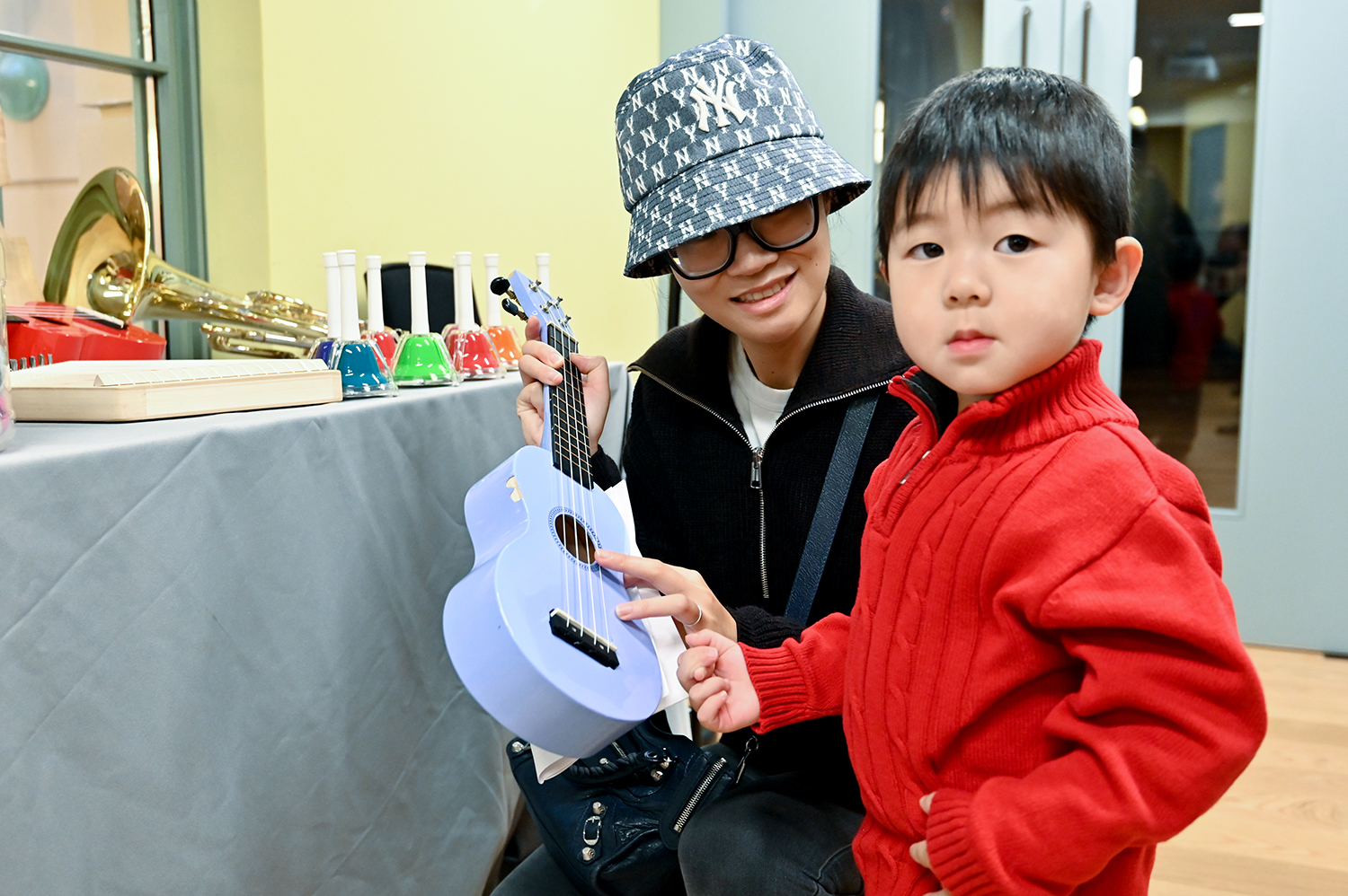 A little boy with black hair and a red jumper poses next to a woman sitting next to him and holding a pale blue ukulele