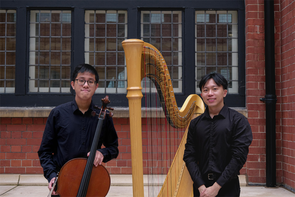 Two male students with dark hair, one holding a cello and the other next to a harp