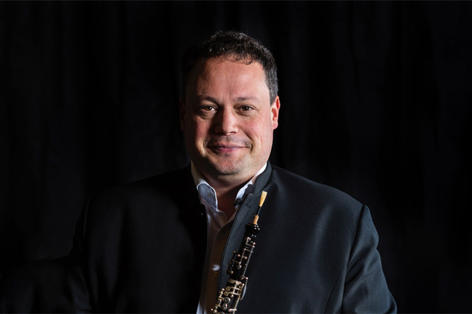 Nick Deutch holds his oboe and smiles in a dark room