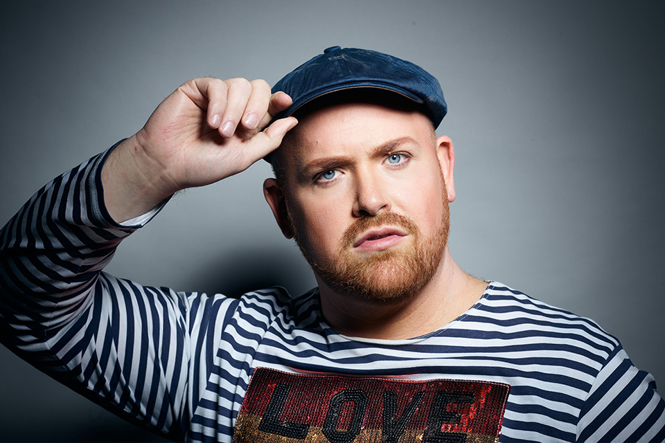 Nicky Spence holds the tip of his flatcap stood against a grey background