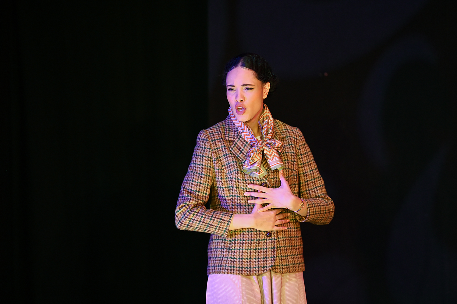 A woman wearing a tweed jacket and scarf singing on stage