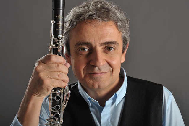 A man with grey hair against a grey backdrop holding a clarinet vertically and smiling at the camera